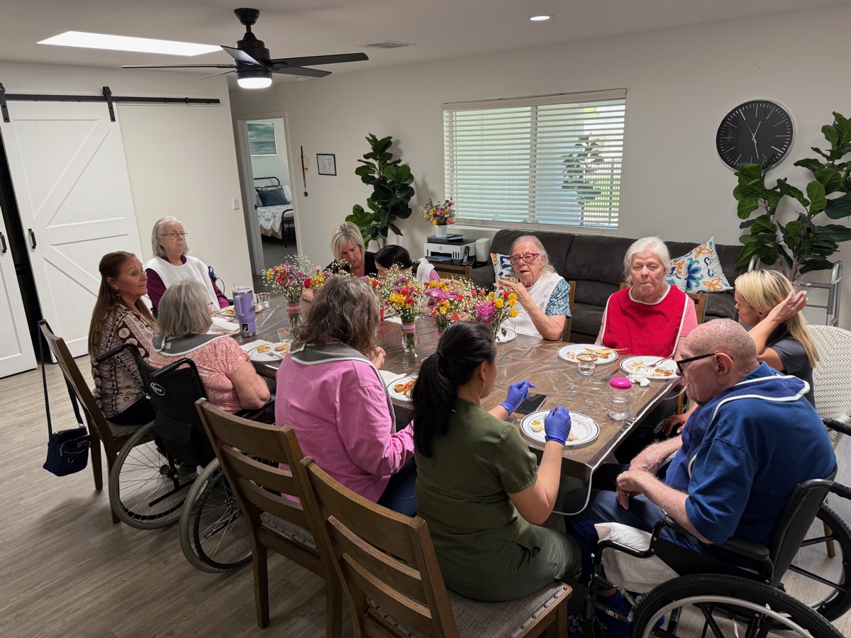 Residents and staff at the dining table chatting over lunch, with bouquets of flowers between them