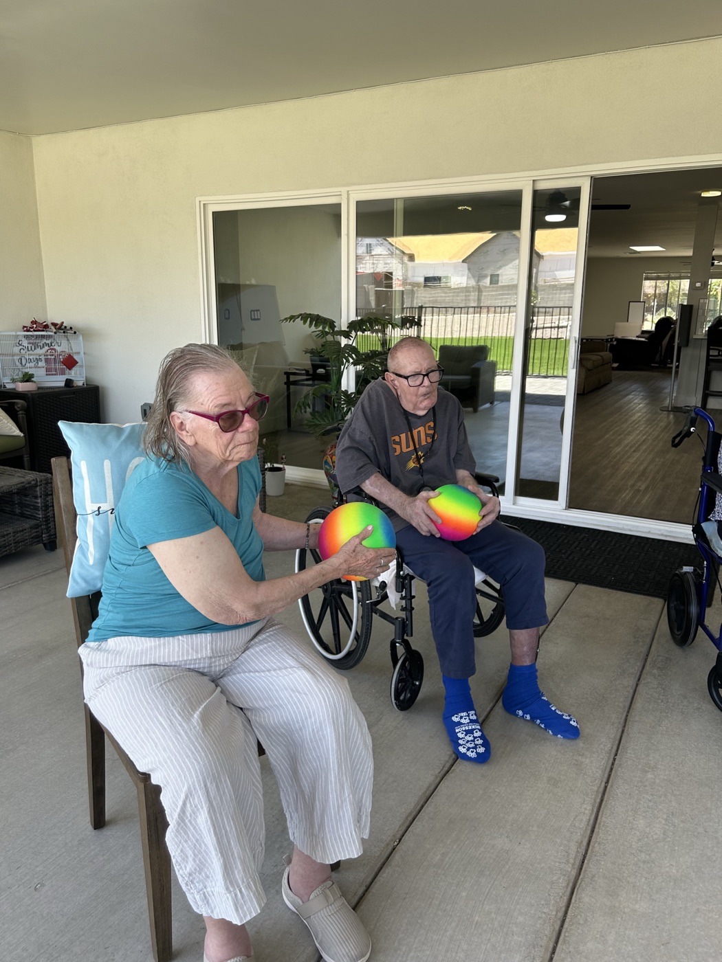 Two residents hold rainbow therapy balls during a seated strength exercise on the patio